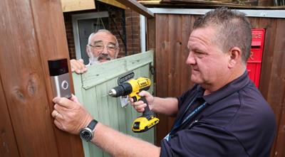 Blue Lamp Trust employee fitting Ring Doorbell on fence door while customer watches from the other side