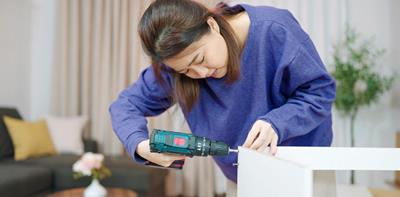 Asian woman in blue top uses a hand held drill on shelf